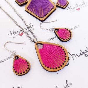 May include: A set of pink and purple thread wrapped wooden earrings and a necklace. The earrings and necklace are shaped like teardrops. The necklace has a silver chain.