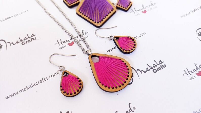 May include: A set of pink and purple thread wrapped wooden earrings and a necklace. The earrings and necklace are shaped like teardrops. The necklace has a silver chain.