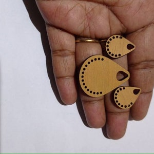 May include: Three wooden teardrop-shaped pendants with a hole at the top and a decorative border of small circles. The pendants are different sizes and are light brown in color.