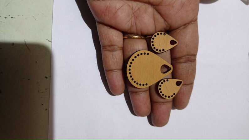 May include: Three wooden teardrop-shaped pendants with a hole at the top and a decorative border of small circles. The pendants are different sizes and are light brown in color.