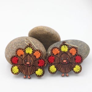 Puede incluir: Un par de pendientes de madera con forma de pavo con plumas de cola bordadas en rojo, naranja y amarillo. Los pendientes tienen ganchos dorados y están colocados sobre un fondo blanco con piedras.