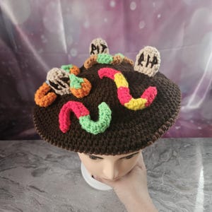 May include: A brown crocheted hat with colorful worms, pumpkins, and tombstones. The hat is designed to look like a graveyard scene, with the worms and pumpkins adding a playful touch. The tombstones have the letters "RIP" and "R.I.P."
