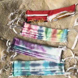 Puede incluir: Cuatro mascarillas faciales con lazos elásticos blancos para las orejas. La mascarilla superior es roja con un patrón blanco y dorado. La siguiente mascarilla es un patrón tie-dye con colores rosa, azul y verde. La tercera mascarilla es un patrón tie-dye con colores morado, azul y blanco. La mascarilla inferior es un patrón tie-dye con colores azul, verde y blanco.