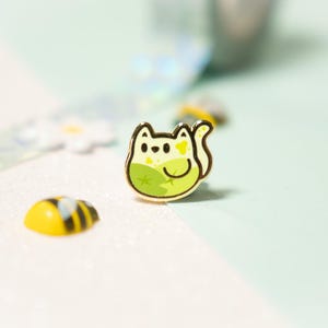Peut inclure: Une épingle en émail dorée représentant un chat de dessin animé avec un motif vert et jaune. Le chat est assis sur une feuille verte et a une fleur jaune sur la tête.