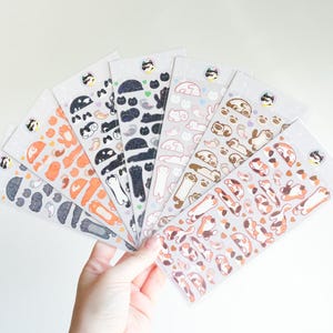 May include: A selection of cat-themed sticker sheets fanned out in a hand. The stickers depict cats in diverse poses and colours, such as black, orange, and brown. Each sheet is adorned with a small cat logo at the top.
