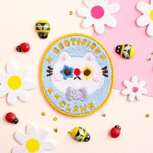 Peut inclure: Un patch brodé bleu et jaune avec un chat blanc portant un nœud papillon violet. Le texte sur le patch est "I CERTIFY I AM A CLOWN".