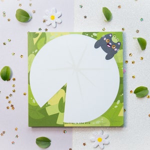 Könnte beinhalten: Ein grünes und weißes Notizbuch mit einer Frosch- und Katzenillustration. Das Notizbuch hat einen weißen Kreis in der Mitte mit dem Text "Sparkles in the Wild" unten.