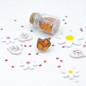 May include: Orange and white enamel pin of a cartoon cat with a surprised expression. The pin is surrounded by small white flowers, leaves, and hearts on a white background.