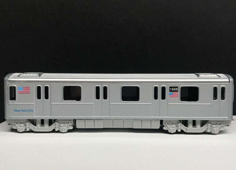 K&ouml;nnte beinhalten: Ein silbernes Spielzeugmodell eines New Yorker U-Bahn-Wagens mit der Nummer 1668 und einer amerikanischen Flagge an der Seite. Der Wagen hat mehrere Fenster und T&uuml;ren. Der Wagen steht auf einer wei&szlig;en Oberfl&auml;che.