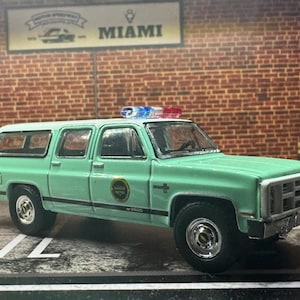 Op de afbeelding: Een mintgroen Chevrolet Suburban model met een politiethema. Het model heeft een lichtbalk op het dak en een "Miami"-bord op de achtergrond. Het voertuig heeft zwarte banden en chromen velgen. Het model heeft een badge aan de zijkant met "Miami Police".