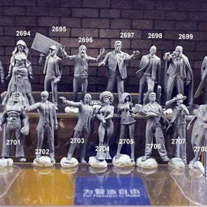 Puede incluir: Una colección de 16 figuras en miniatura pintadas de gris, cada una con una pose y una expresión únicas. Las figuras están dispuestas en dos filas sobre un expositor transparente. El expositor tiene una base negra y una etiqueta blanca con el texto "Para la libertad de hacer".