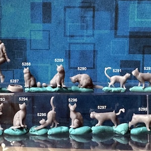 May include: A collection of small, gray cat figurines in various poses, displayed on a clear shelf against a blue background. The figurines include cats sitting, stretching, and interacting. A couple figurine is also present.