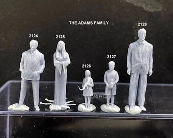 May include: A collection of gray miniature figurines representing the Adams Family. The figures include a man in a suit, a woman in a long dress, a girl, a boy, and another man in a suit. The text "THE ADAMS FAMILY" is above the figures.