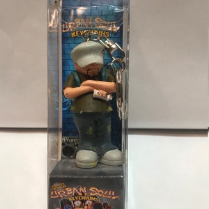 Può includere: Un portachiavi in plastica con un personaggio dei cartoni animati che indossa una camicia marrone, jeans blu e un berretto da baseball bianco. Il personaggio tiene una bomboletta spray in una mano e ha un design a spruzzo di vernice sui suoi vestiti. Il portachiavi è confezionato in una scatola di plastica trasparente con le parole "Urban Soul Keychains" stampate sul davanti.