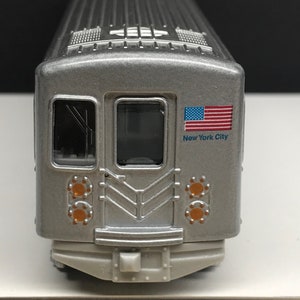 K&ouml;nnte beinhalten: Ein silbernes Spielzeugmodell eines New Yorker U-Bahn-Zuges mit einer amerikanischen Flagge und dem Schriftzug "New York City" an der Seite.