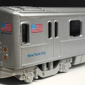 K&ouml;nnte beinhalten: Ein silbernes Spielzeugmodell eines New Yorker U-Bahn-Wagens mit zwei amerikanischen Flaggen und dem Schriftzug "New York City" an der Seite.
