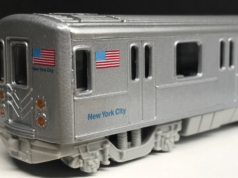 K&ouml;nnte beinhalten: Ein silbernes Spielzeugmodell eines New Yorker U-Bahn-Wagens mit zwei amerikanischen Flaggen und dem Schriftzug "New York City" an der Seite.