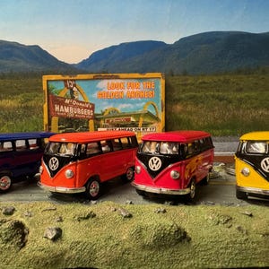 May include: Four miniature Volkswagen bus keychains in different colors: blue, red, pink, and yellow. Each keychain has a black VW logo and a black window frame. The keychains are arranged in a row on a surface with a vintage McDonald's sign in the background.
