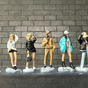 以下が含まれることがあります： さまざまなファッショナブルな服装をした6人のミニチュアフィギュア。小さな岩のような台座の上に立っています。フィギュアは、黄色のジャケット、白いトップス、黒いジャケット、タンのトラックスーツ、ターコイズのジャケット、タンの帽子が付いた黒いジャケットなど、さまざまなスタイルで着飾っています。