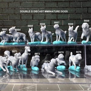 May include: A collection of miniature diecast dog figurines in various poses, displayed on a clear shelf against a brick background. Each dog is a light gray color and rests on a small, blue base. The text "DOUBLE G DIECAST MMINIATURE DOGS" is at the top.