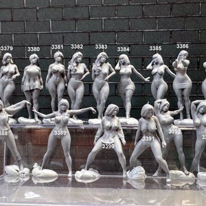 Peut inclure: Une collection de 14 figurines grises de femmes dans différentes poses. Les figurines sont disposées en deux rangées sur un présentoir transparent. Chaque figurine porte un numéro imprimé sur sa base.