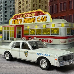 Puede incluir: Un coche de policía blanco con una señal auxiliar azul y blanca, estacionado frente a un restaurante rojo y blanco con un letrero que dice "Mike's Dining Car".