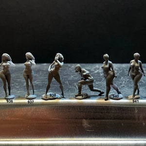 May include: A set of ten grey figurines, each standing on a small base. The figurines are arranged in a row on a silver surface. The figurines are all different, but they all have a similar style. Some of the figurines are wearing clothing, while others are nude.