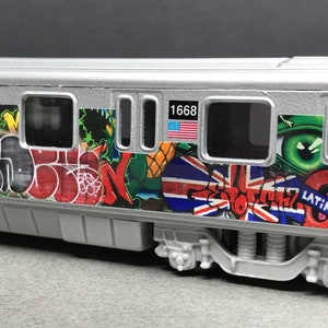 Puede incluir: Un vagón de tren de juguete plateado con arte de graffiti en el lateral. El vagón tiene el número "1668" y una bandera estadounidense en el lateral. El arte de graffiti incluye una Union Jack, un ojo verde y la palabra "LATINOS".