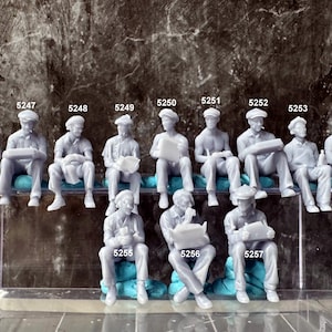 May include: A collection of miniature gray figurines of seated men, each with a hat and various props like books and a bottle. The figures are arranged in rows, with numbers above and below them, suggesting a model kit or diorama.