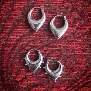 Puede incluir: Cuatro pendientes de aro de plata con diferentes diseños. Dos pendientes tienen forma de lágrima puntiaguda, y dos pendientes tienen forma de lágrima puntiaguda con pequeños pinchos alrededor del borde.