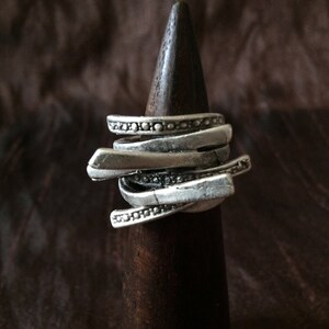 Könnte beinhalten: Ein silberner Ring mit mehreren Bändern, jedes mit einem anderen Design, auf einem hölzernen Ringständer ausgestellt.