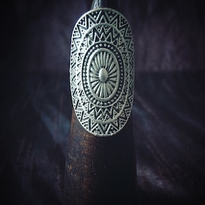 Könnte beinhalten: Ein silberner Ring mit geometrischem Design. Der Ring ist oval geformt und hat einen zentralen Kreis mit einem Sonnenstrahlenmuster. Der Ring befindet sich auf einem Holzständer.