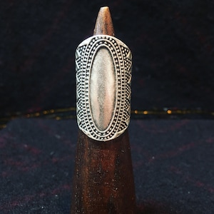 Könnte beinhalten: Ein silberner Ring mit einem ovalen Zentrum und einem geometrischen Design an den Rändern. Der Ring wird auf einem hölzernen Ringständer präsentiert.