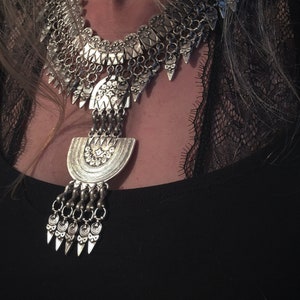 Può includere: Una collana di dichiarazione argentata con un grande ciondolo decorato. Il ciondolo ha la forma di una mezzaluna e presenta dettagli intricati. La collana è realizzata con catene di metallo e ha un design a strati.