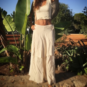 Può includere: Una gonna bianca avvolgente con un design a strati. La gonna è realizzata in un tessuto morbido e fluido e ha una vestibilità rilassata. La gonna è indossata da una persona in piedi in un ambiente da giardino.