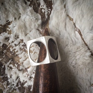 May include: A silver ring with a geometric design. The ring is shaped like a cube with a circular cutout on each side. The ring is sitting on a wooden surface.