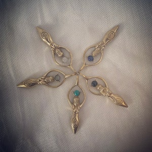 K&ouml;nnte beinhalten: F&uuml;nf goldfarbene Metallanh&auml;nger in Form einer Frau, die einen Kreis h&auml;lt. Jeder Anh&auml;nger hat einen andersfarbigen Edelstein im Kreis. Die Anh&auml;nger sind sternf&ouml;rmig angeordnet.