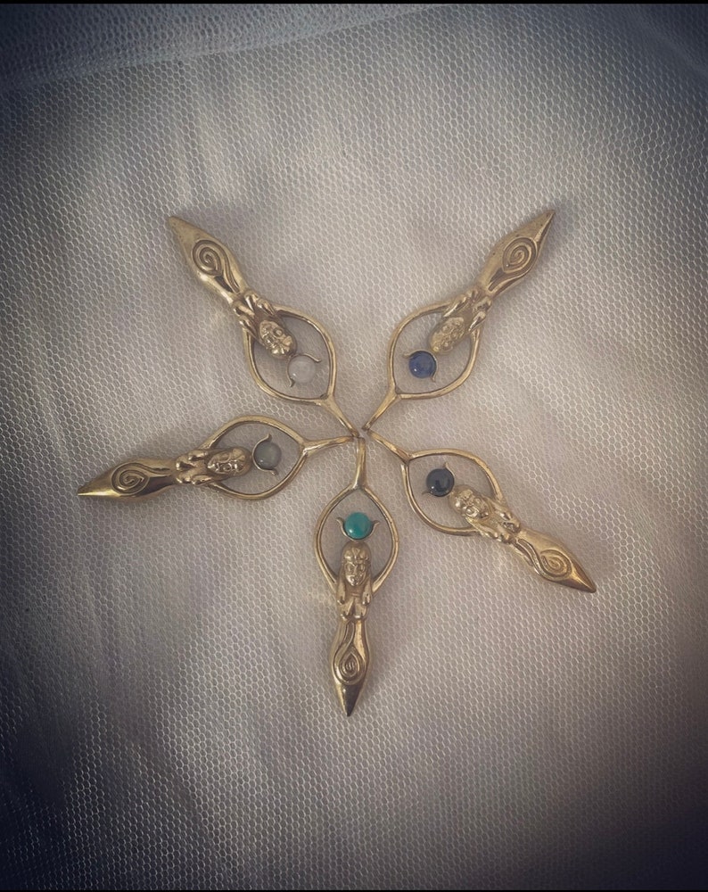 K&ouml;nnte beinhalten: F&uuml;nf goldfarbene Metallanh&auml;nger in Form einer Frau, die einen Kreis h&auml;lt. Jeder Anh&auml;nger hat einen andersfarbigen Edelstein im Kreis. Die Anh&auml;nger sind sternf&ouml;rmig angeordnet.