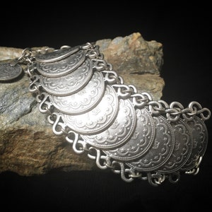 Puede incluir: Un brazalete de plata con una cadena y múltiples dijes en forma de moneda. Las monedas tienen diseños intrincados y están conectadas a la cadena con bucles.