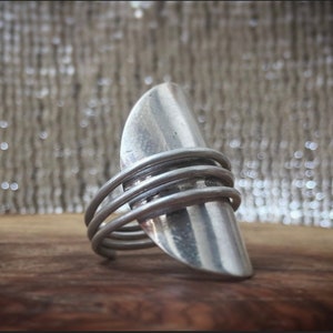 Op de afbeelding: Een zilveren ring met een brede, gebogen band en meerdere dunne banden eromheen gewikkeld.