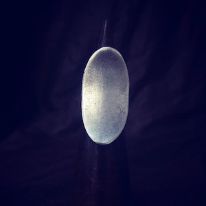 Può includere: Un anello ovale in argento con una superficie liscia e leggermente strutturata. L'anello è esposto su un supporto grigio scuro.