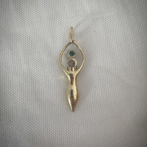 K&ouml;nnte beinhalten: Ein goldener Anh&auml;nger mit einer stilisierten Frauenfigur, &uuml;ber deren Kopf sich ein Augensymbol befindet. Das Auge ist mit einem gr&uuml;nen Edelstein besetzt.