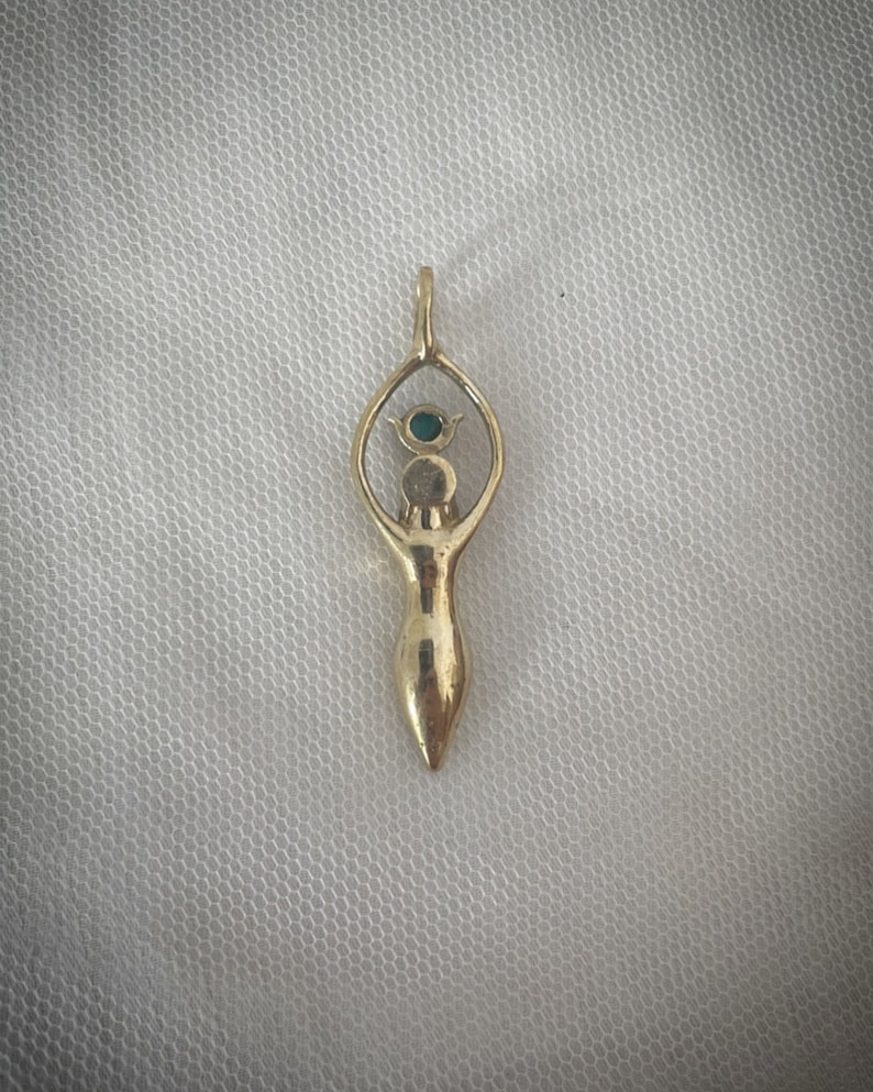 K&ouml;nnte beinhalten: Ein goldener Anh&auml;nger mit einer stilisierten Frauenfigur, &uuml;ber deren Kopf sich ein Augensymbol befindet. Das Auge ist mit einem gr&uuml;nen Edelstein besetzt.