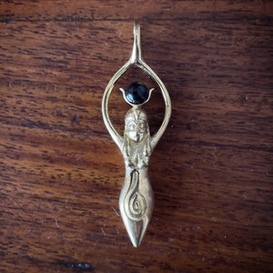 K&ouml;nnte beinhalten: Ein goldener Anh&auml;nger mit einer stilisierten weiblichen Figur, die oben einen schwarzen Edelstein tr&auml;gt. Die Figur steht mit erhobenen Armen und einem Spiralmuster am unteren K&ouml;rper.