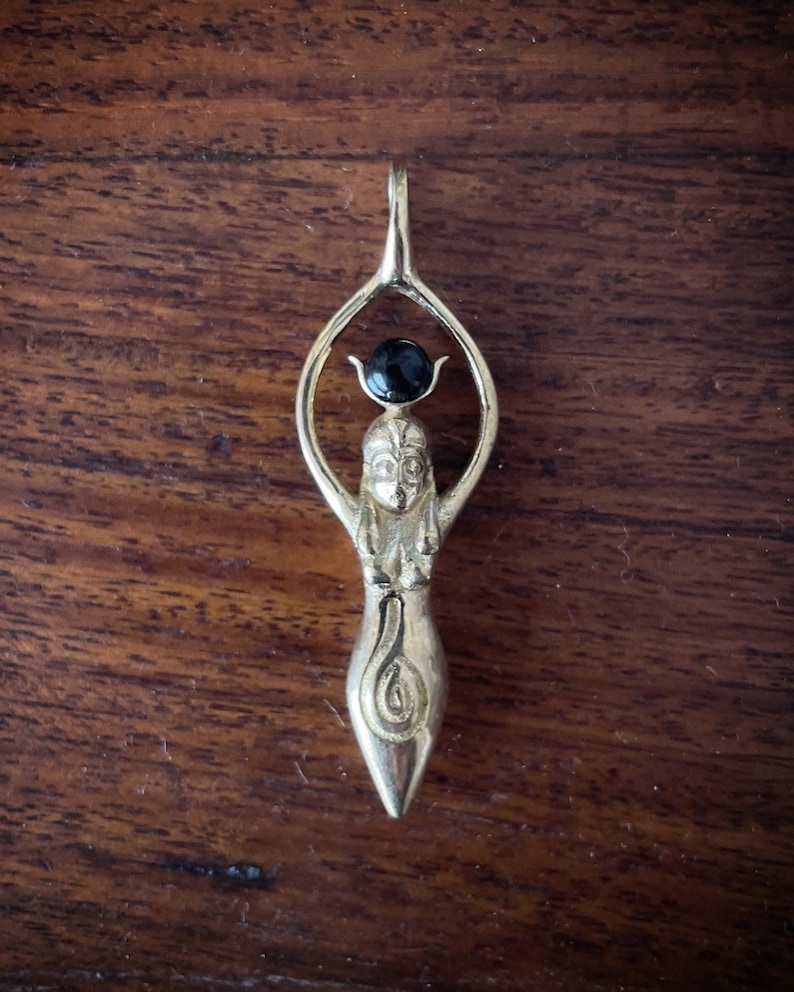 K&ouml;nnte beinhalten: Ein goldener Anh&auml;nger mit einer stilisierten weiblichen Figur, die oben einen schwarzen Edelstein tr&auml;gt. Die Figur steht mit erhobenen Armen und einem Spiralmuster am unteren K&ouml;rper.