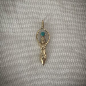 K&ouml;nnte beinhalten: Ein goldener Anh&auml;nger mit einer stilisierten Frauenfigur und einem t&uuml;rkisfarbenen Edelstein an der Spitze. Der Anh&auml;nger hat die Form einer Tr&auml;ne und ist detailliert gestaltet.