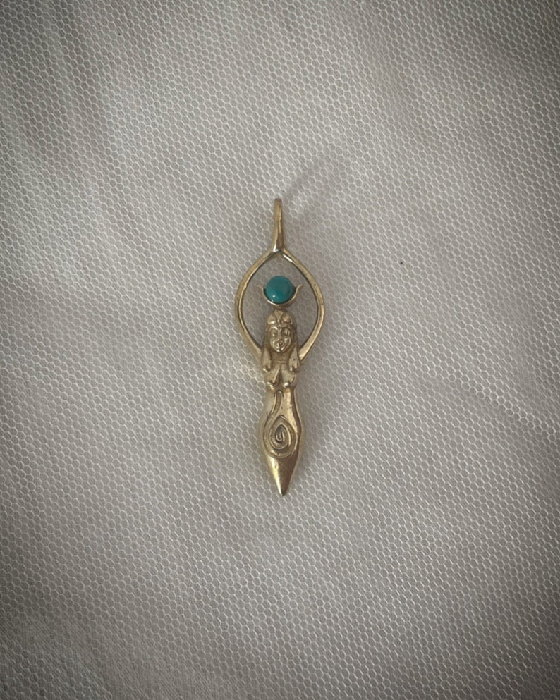 K&ouml;nnte beinhalten: Ein goldener Anh&auml;nger mit einer stilisierten Frauenfigur und einem t&uuml;rkisfarbenen Edelstein an der Spitze. Der Anh&auml;nger hat die Form einer Tr&auml;ne und ist detailliert gestaltet.