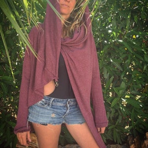 May include: A burgundy hooded wrap sweater with long sleeves and a draped front. The sweater is paired with denim shorts. The background features green foliage.