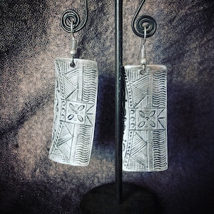 Peut inclure: Une paire de boucles d'oreilles rectangulaires argentées avec des motifs géométriques complexes. Les boucles d'oreilles présentent un mélange de lignes, de triangles et de motifs floraux, suspendues à de simples crochets. Fond sombre.