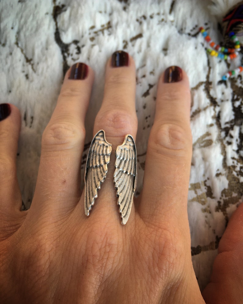 May include: A silver-toned ring featuring a split wing design. The ring is worn on a finger, with the wings extending outwards. The ring's details include feather-like textures and a polished finish. The background is a textured, off-white surface.